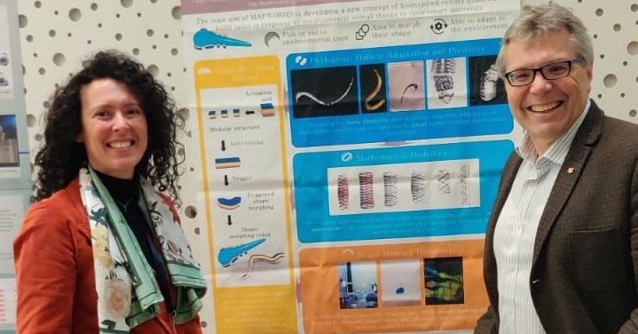 Professor Arianna Menciassi standing beside the MAPWORMS poster exposed at ERF2023, an event in the field of robotics, together with a participant of the exhibition